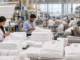 Workers in a towel manufacturing plant folding and stacking white towels at a production station the size of a large factory floor